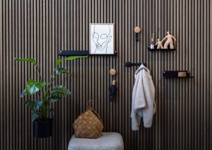WoodUpp-Wandverkleidung aus schlanken, senkrechten Holzlamellen, kombiniert mit WoodUpp runden Holzklebehaken, schwarzen Metall-Regalborden und einem Pflanzenhalter. Schwarze Metallakzente mit sichtbaren goldfarbenen Schrauben betonen das Design. Auf den Regalen stehen Rahmen, Duftstäbchen und Tonfiguren; an den Haken hängen Mantel und Schirm.