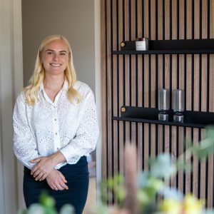 Porträt einer lächelnden Frau neben einer Holzlamellenwand mit zwei schwarzen WoodUpp-Wandregalen. Die schlanken, matt schwarzen Metallregale zeigen goldene Befestigungsknöpfe; darauf eine Duftkerze (No. 02) und zwei gerippte Gläser. Die Frau trägt eine weiße Lochstickerei-Bluse und dunklen Rock.