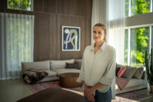 Eine lächelnde Frau im hellen Hemd steht im Vordergrund des Bildes, warmes Tageslicht fällt durchs Fenster. Im Hintergrund WoodUpp Lamellen-Wandverkleidung aus vertikalen Holzlamellen und ein runder WoodUpp Couchtisch aus warmem Holz, dazu helles Ecksofa mit Kissen, Plaid, Teppich, Kaktus und luftige Vorhänge.