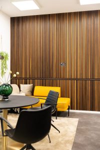WoodUpp Holzlamellen-Wandpaneele dominieren das Bild: vertikale, schlanke Holzleisten im warmen Holzton mit feiner Maserung und kontrastierenden schwarzen Fugen sowie zusätzlicher horizontaler Blende. Davor modernes Sitzarrangement mit gelber Bank, schwarzen Stühlen, rundem Tisch, Teppich und grüner Dekovase in hellem, minimalistischem Interieur.