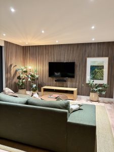 Modernes Wohnzimmer mit grünem Sofa, runder Couchtisch und an der Wand montiertem Fernseher. Warm beleuchtete WoodUpp-Lamellenwand aus senkrechten, engstehenden Holzleisten in heller Eichenoptik als Akzent, schlichtes Holz-Lowboard, zwei große Zimmerpflanzen, Juteteppich und gerahmtes Landschaftsbild rechts; weiches Deckenlicht, gedämpfte Farben.