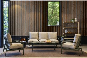 WoodUpp Lounge-Ecke mit dreisitzigem Sofa und zwei gepolsterten Sesseln in olivegrünem Metallrahmen und hellbeigen Leinenkissen, niedrigem Glastisch mit Tablett und Gläsern sowie rundem Beistelltisch. Dazu offenes WoodUpp-Regal aus hellem Holz mit Pflanzen und Deko, vor Holzlamellenwand und Fenster.