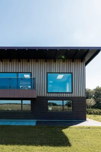 Modernes zweigeschossiges Haus mit WoodUpp-Holzlamellen-Fassade im Obergeschoss: schmale, vertikal angeordnete helle Holzleisten als Fassadenverkleidung. Sichtbar sind außerdem ein WoodUpp-Holzbalkonrand mit Glasgeländer sowie große Fenster mit bläulicher Verglasung, darunter dunkle Ziegelwand und grüner Rasen. Die Lamellen zeigen gleichmäßigen Abstand und feine Holzstruktur, das Dach mit breitem Vorsprung wirft Schatten.