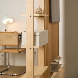 WoodUpp Raumteiler/Regalsystem aus hellen Eichenleisten mit modularen Holzablagen, darauf eine Garnrolle. Eingehängte WoodUpp Metallbox in creme/beige als geschlossene Aufbewahrung und eine schmale WoodUpp Metallablage mit Fernbedienungen sichtbar. Elegante, minimalistische Optik mit sichtbaren Metallverschraubungen; im wohnlichen Hintergrund Schreibtisch, Stuhl und Lampe, verschwommen.