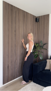 Eine lächelnde Frau steht vor einer Wand, die mit WoodUpp Akustikpaneelen in Nussbaum-Optik verkleidet ist. Die Paneele sind über Eck installiert und verleihen dem Raum eine moderne, warme Ausstrahlung. Eine große Pflanze und ein dunkles Sofa ergänzen die Szene.