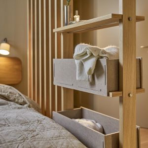 WoodUpp Raumteiler-Regal aus Eichenholz mit senkrechten Lamellen, zwei grauen Filz-Aufbewahrungsboxen, eine mit zusammengerolltem Strickpullover, sowie einer schmalen Ablage mit Vase und Flakon. Massive Holzkonstruktion mit sichtbaren Messing-Schrauben neben Bett und Leselampe. Die Filzboxen stecken in Holzschienen und betonen warmes, neutrales Schlafzimmerambiente.