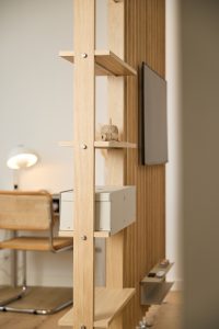 WoodUpp-Raumteiler und Regalsystem aus hellem Eichenholz mit senkrechten Lamellen, sichtbaren Metallbolzen, natürlicher Holzmaserung, mehreren schwebenden Ablageböden, einer beigefarbenen Schubkastenbox von WoodUpp sowie einer kleinen Dekoration mit Garnrolle. An der Trennwand hängt ein Flachbildschirm; im Hintergrund Büroecke mit Flechtstuhl und Schreibtischlampe.