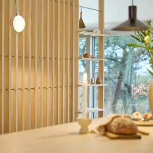 Helles Interieur mit WoodUpp Lamellenwand aus Eichenholz, versehen mit schlanken Metallhaken und integrierten WoodUpp Regalböden als luftige Fachregale, dekoriert mit kleinen Schalen und Dosen. Im Vordergrund brennende Kerze und Brot, im Hintergrund große Fenster mit Blick ins grüne Gartengelände und warmem Holzton.