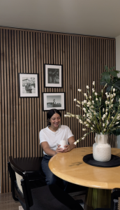 Eine Frau genießt ein Getränk an einem runden Tisch, hinter ihr eine markante Wandgestaltung mit WoodUpp Akustikpaneelen. Diese Paneele zeigen dunkle Holzlamellen, vermutlich in Nussbaum, montiert auf schwarzem akustischem Filz. Die warme Holzwand ist mit drei gerahmten Bildern dekoriert und schafft eine gemütliche Essbereich-Atmosphäre.