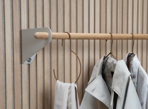 Bild zeigt WoodUpp Garderobenlösung: horizontale Eichenstange gehalten von matten, dreieckigen Metall-Wandhalterungen, montiert an einer lamellenartigen Eichenholzverkleidung. Darauf Lederbügel und ein offener Lederring für Schals sowie zwei helle Mäntel/Trenchcoats aufgehängt, klarer, skandinavisch-minimaler Stil. Die warme, naturfarbene Eiche des Rohrs harmoniert mit sandfarbenen Mänteln und cognacfarbenen Lederhängern.