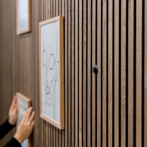 Nahaufnahme einer WoodUpp Lamellenwand in warmem Eichenfarbton aus hellbraunen, feinstrukturierten Holzleisten mit gleichmäßig montierten Vertikalschluchten, bestückt mit einem kleinen runden schwarzen WoodUpp Wandhaken. Eine Hand hängt zwei filigrane Holzrahmen mit minimalistischen Linienzeichnungen an die Lamellen, eine sachliche, elegante Präsentation im Innenraum.