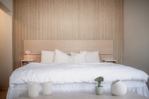 Minimalistisches Schlafzimmer mit WoodUpp-Produkten: helles Holz-Kopfteil, senkrechte WoodUpp-Lamellenwand in Eichenoptik und schmale WoodUpp-Nachttische. Naturbelassene, helle Holzoberfläche, klare Linien, kombiniert mit weißer Bettwäsche, Kissen, Lampe, Kerzen und Topfpflanze, mit feinen Schattenfugen und warmem Ambiente.