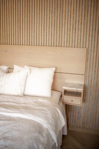 WoodUpp: Ruhiges Schlafzimmer mit hellen Eichenholz-Lamellen an der Wand, passendem Holz-Kopfteil und einem schwebenden WoodUpp-Nachttisch mit Marmorplatte, offener Ablage und runder Schubladengriff. Die Oberfläche zeigt feine Maserung und natürliche, helle Töne. Davor liegt ein weißes Bett mit strukturierten Kissen und weicher Leinenbettwäsche im minimalistischen, skandinavischen Stil.