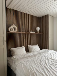 Schlafzimmer mit dunkelbrauner WoodUpp-Holzlamellen‑Wand, darauf ein schlankes WoodUpp‑Schweberegal mit Keramik‑Dekovasen, zwei Metall‑Kerzenhaltern, einer kleinen Glaskugel und einer runden Tischleuchte. Weiße Bettwäsche und Kissen im Vordergrund betonen die warme, minimalistische Atmosphäre. Die WoodUpp-Produkte zeigen feine Maserung, filigrane Schattenfugen und warmen Holzton.