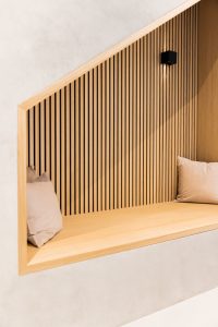 WoodUpp: Eingelassene Sitznische mit heller Holz-Sitzbank und vertikaler WoodUpp-Lamellenwand aus schlanken Eichenlatten, zwei beigefarbenen Kissen und einer kleinen schwarzen Wandlampe. Warmer Naturton, präzise Nut- und Feder-Optik, filigrane Fugen und abgeschrägter Holzrahmen schaffen ruhige, minimalistische Atmosphäre sowie elegantes, zeitloses Design und hochwertige Handwerksqualität.