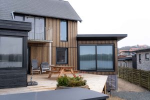 Wohnhaus mit WoodUpp-Fassade aus schmalen, vertikalen Holzlamellen und großen Fenstern, ergänzt durch helle WoodUpp-Terrassendielen. Auf der Terrasse stehen ein Picknicktisch, zwei Klappstühle und ein Kübel mit Nadelgehölz; dunkle Fensterrahmen und eine Schiebetür spiegeln den See im Hintergrund, moderner Anbau mit Flachdach und Metallgeländer am Balkon.