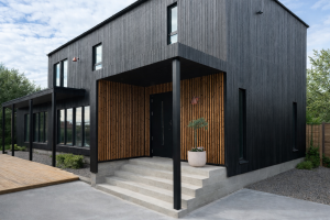 Dieses Bild zeigt ein modernes Haus mit einer dunklen Fassade. Der zurückgesetzte Eingangsbereich ist das Highlight, verkleidet mit WoodUpp Akustikpaneelen in einer warmen Holzoptik, wahrscheinlich Eiche. Die vertikalen Lamellen schaffen einen schönen Kontrast zur schwarzen Umgebung und der Betontreppe.